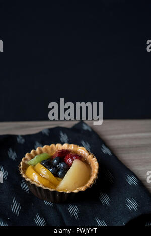 Kleiner gemischter Obstkuchen auf dunklen Serviette auf hellem Holz vor der dunklen Wand Stockfoto