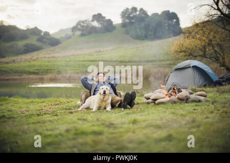 Mitte der erwachsenen Mann zu entspannen und die Arme hinter seinem Kopf neben seinen Hund auf einem Campingplatz in der Nähe eines Flusses. Stockfoto