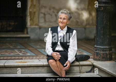 Ältere Erwachsene Frau sitzt auf einem Schritt mit den Händen auf den Knien in einem Foyer. Stockfoto