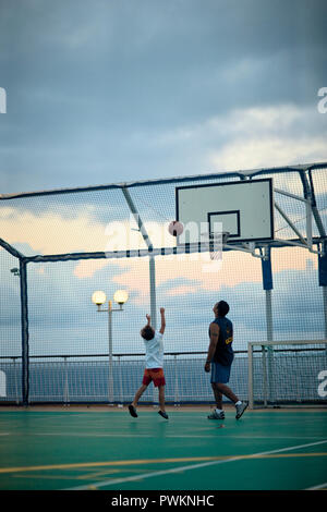 Man beobachtet, wie ein Junge einen Basketball in den Ring wirft auf einem eingezäunten seaside Basketballplatz. Stockfoto