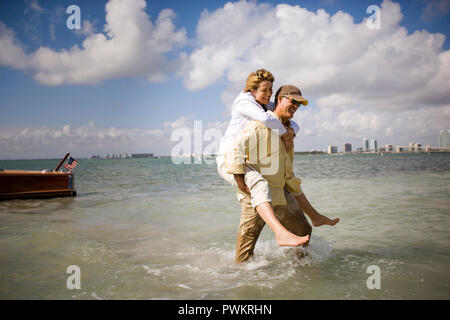 Lächelnd reifer Mann huckepack seine Frau von einem Boot zum Ufer am Strand. Stockfoto