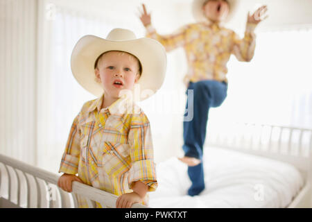 Porträt eines Jungen einen Hut tragen, beim Stehen auf einem Bett mit seinem älteren Bruder. Stockfoto