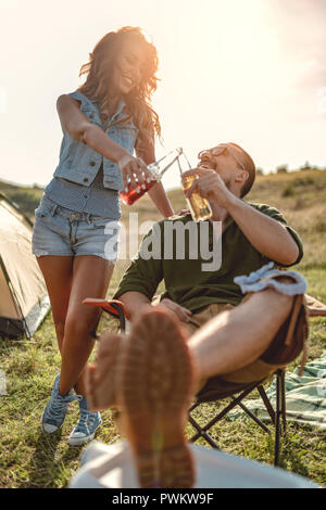 Glückliche junge Paar genießt eine sonnige Tag in der Natur. Sie klirren Bierflaschen vor einem Campingplatz Zelt. Stockfoto