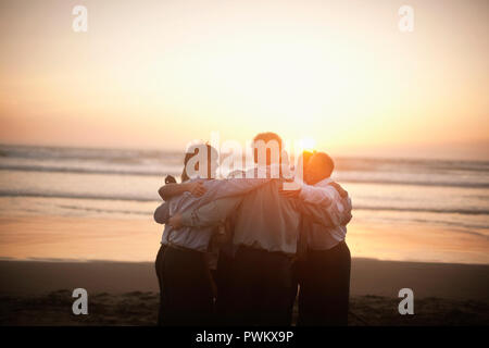 Kleine Gruppe von Geschäftsleuten stehen in einem Kreis und einander umarmen, während sie am Strand bei Sonnenuntergang. Stockfoto
