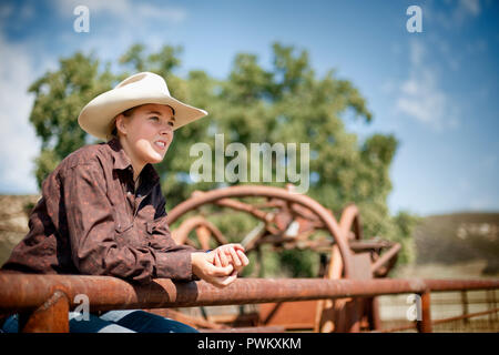 Teenage Mädchen mit einem Cowboyhut sitzt zwischen den rostigen Geländer einer Corral und schaut weg nachdenklich, als sie posiert für ein Portrait. Stockfoto