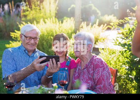 Im Sommer, eine Familie von drei Generationen, die um einen Tisch im Garten versammelt, um eine Mahlzeit zu teilen. Großeltern teilen ein Video mit Ihren kleinen daugh