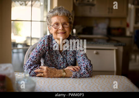 Porträt einer fröhlichen älteren Frau alleine sitzen an ihrem Küchentisch. Stockfoto