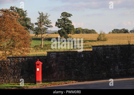 Die Sonne scheint auf eine helle rote Britische Post Box stand vor einer dunklen Mauer aus Stein, mit bepflanzten Felder und englischen Landschaft im Hintergrund. Stockfoto