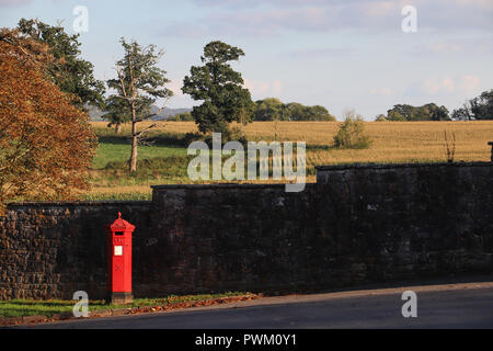 Die Sonne scheint auf eine helle rote Britische Post Box stand vor einer dunklen Mauer aus Stein, mit bepflanzten Felder und englischen Landschaft im Hintergrund. Stockfoto