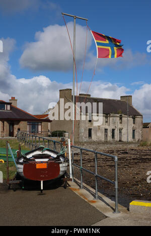 Ein sehr zerschlissenen Fahne von Orkney, Schottland, Fliegen im starken Wind in vom Pier in St. Mary's. Strahlend blauen Himmel, weiße Wolken, Boot, Gebäude, Ufer. Stockfoto