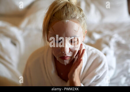 Junge Frau mit weißem Papier Maske beim Sitzen auf dem Bett zu Hause. Stockfoto