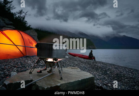 Tee und Kaffee mit einem Gasherd auf dem Campingplatz mit roten neigen, lit. Blick auf See bei bewölkt Nacht. Unscharfe Silhouette eines Mannes auf Hintergrund. Stockfoto