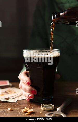 Flasche Guinness in einem großen Glas durch eine Hand mit rotem Nagellack auf einen hölzernen Tisch gehalten wird gegossen umgibt in der Flasche top, Flasche op Stockfoto