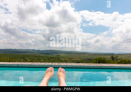 Eine Nahaufnahme der Frauen Füße mit einem zauberhaften Blick auf einen glasklaren Pool und großen afrikanischen Busch in die Berge führen Stockfoto