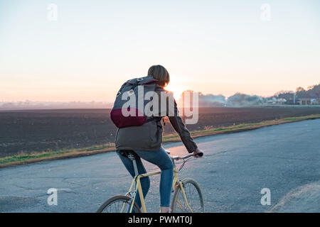 Weibliche Pendler mit dem Fahrrad aus der Stadt in einen Vorort. Junge Frau geht nach Hause mit dem Fahrrad von der Arbeit entlang der Straße bei Sonnenuntergang Stockfoto