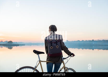 Weibliche Radfahrer geniessen Sie einen wunderschönen blauen Stunde Szene am See. Frau steht mit Bike und sieht am schönen See und Sonnenuntergang Stockfoto