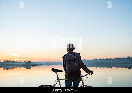 Weibliche Radfahrer geniessen Sie einen wunderschönen blauen Stunde Szene am See. Frau steht mit Bike und sieht am schönen See und Sonnenuntergang Stockfoto