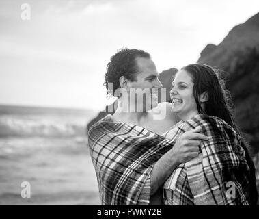 Lächelndes Paar in eine Decke eingewickelt nach dem Schwimmen im Wasser am Strand. Stockfoto