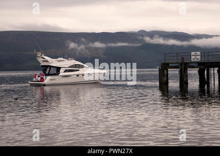 Motor Cruiser nähern Luss Pier am Loch Lomond am frühen Morgen mit Nebel auf dem Hügel dahinter. Stockfoto