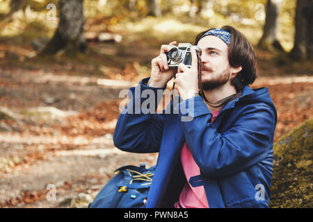 Horizontale Seitenansicht Portrait von junge Bartgeier männliche Touristen tragen rosa t-shirt, blau Mantel und Jeans mit Rucksack und Kamera, wunderschöne Sonnenuntergang auf Moss vor Baum umgeben von Wald, Reisen Lifestyle erfolg konzept Abenteuer Sommer aktiv Urlaub Outdoor bergsteigen Sport Stockfoto