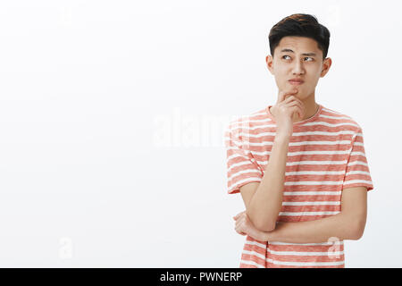 Portrait von gestörten jungen Asiaten versuchen, Plan oder Idee denken, stehen in nachdenklicher Pose mit der Hand am Kinn, in Frage gestellt und an der oberen linken Ecke zögerlich, die Annahmen Stockfoto