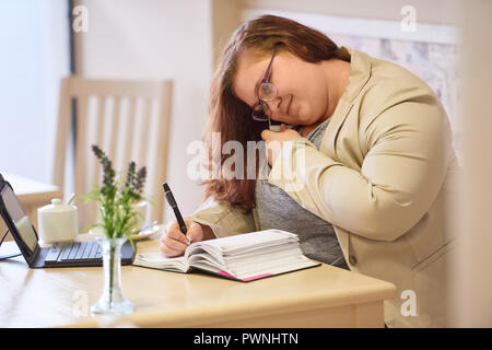 Übergrößen Frau besetzt Multitasking im Café, im Gespräch in auf ihrem Mobiltelefon, während die, die sich Notizen in ihrem Tagebuch, während Sie Ihren Kalender auf der Suche nach c Stockfoto