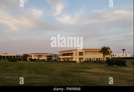 Palmen und anderen angelegten Bäume auf dem Hintergrund einer Resort Hotel in Ägypten, Sharm el-Sheikh bei Sonnenuntergang. Schönen niedrigen eins-zwei-stöckige Häuser - vil Stockfoto