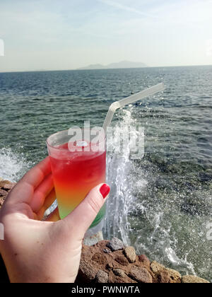Ein Glas mit einem farbigen puff Cocktail in weiblicher Hand vor dem Hintergrund der starken Wellen des Meeres. Stockfoto