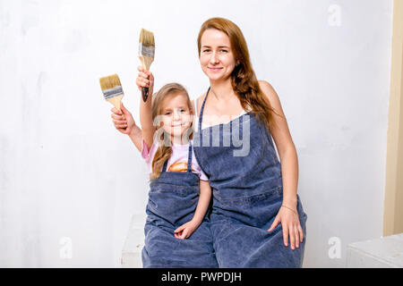 Reparatur in der Wohnung. Glückliche Familie, Mutter und Tochter in Schürzen vorbereitet die Wand mit weißer Farbe zu malen. Mit Quasten in ihren Händen sitzen und Lächeln Stockfoto