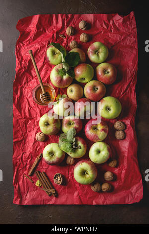 Reif Organic Gardening grün rote Äpfel mit Blätter, Nüsse, Zimt und Glas Honig auf Rot hell zerknittertes Papier über dunkle Textur Hintergrund. Flat l Stockfoto