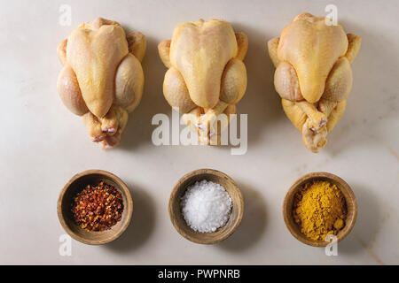 Drei rohe organische ungekocht ganz gelb Mais mini Huhn in der Zeile mit Salz, Kurkuma Pulver, red hot chili Pfeffer in Schalen über Marmor Hintergrund. Coo Stockfoto