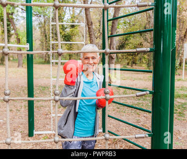 Portrait von Happy Passen ältere Frau mit Boxhandschuh im Fitnessbereich im Freien in Sportbekleidung. Aktive alte Menschen Fitness Lifestyle Stockfoto