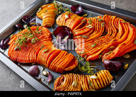 In dünne Scheiben geschnitten Hälften der Butternut Kürbis gebacken mit roten Zwiebeln, frischem Thymian, Kürbiskerne und Gewürze auf einem Backblech auf einer konkreten Tabelle, Ansicht von Stockfoto