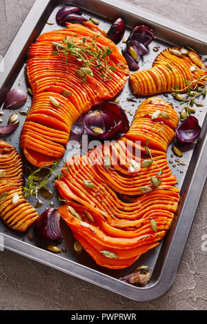 In dünne Scheiben geschnitten Hälften der Butternut Kürbis gebacken mit roten Zwiebeln, frischem Thymian, Kürbiskerne und Gewürze auf einem Backblech auf einer konkreten Tabelle, vertikale v Stockfoto