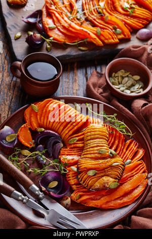 In dünne Scheiben geschnitten geröstet Hälften der Butternut Kürbis mit roten Zwiebeln, Thymian, Kürbiskerne und Gewürze auf einem Steingut Teller auf einen alten Holztisch mit k Stockfoto