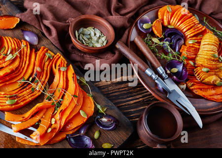 Gebratene Hälften der Butternut Squash oder Kürbis mit roten Zwiebeln, Thymian, Kürbiskerne und Gewürze auf einem Steingut Teller auf einen alten Holztisch wit in Scheiben geschnitten Stockfoto