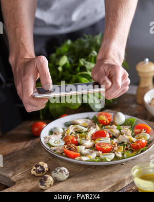 Appetitlich gesund Salat aus biologischen Zutaten in einem Teller mit mens Hand das Foto von Smartphon auf einem Holztisch. Stockfoto