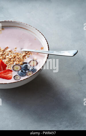 Weiße Schüssel mit hausgemachtem Müsli, in Scheiben geschnittene Erdbeeren, Joghurt, Blaubeere, Löffel - natürliche Frühstück auf einem grauen Tabelle. Stockfoto
