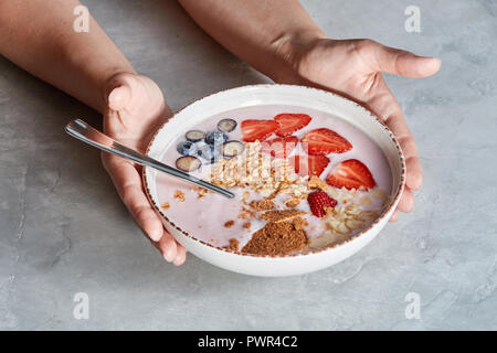 Frau Hände halten Schüssel mit appetitlich gesund Müsli Nachtisch mit Stücken von Erdbeeren, Sahne, Haferflocken auf grauem Beton Hintergrund Stockfoto