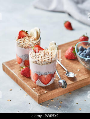 Müsli, Hälften von Erdbeeren, Banane, Chia Samen mit Joghurt in Gläsern auf einem Holzbrett auf grauem Beton, Platz für Text. Stockfoto