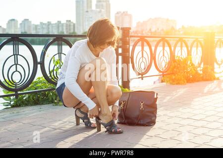 Outdoor Sommer Portrait von reife Frau in Sonnenbrille mit einer Tasche und Schuhe am Abend sonnig Stadt. Stockfoto