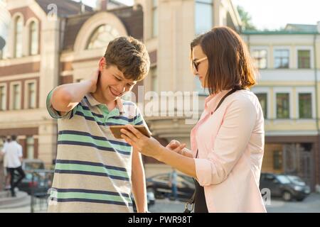 Eltern und Teenager, Beziehung. Die Mutter ihrem Sohn etwas in das Handy zeigt, junge peinlich ist, lächelnd, seine Hände auf seinen Kopf, cit Stockfoto