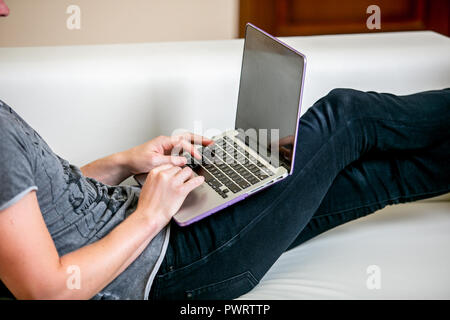 Junger Mann mit Brille, der an einem Notebook arbeitet in einem Home Office konzentriert. Druckt auf der Tastatur und scrollt Text auf dem Bildschirm beim Liegen auf dem Sofa, close-up Stockfoto