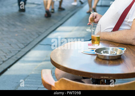 Ein Mann mit großen Bauch sittiing draußen in einer Bar mit einem Arm auf dem runden Holztisch Bier trinkt. Stockfoto