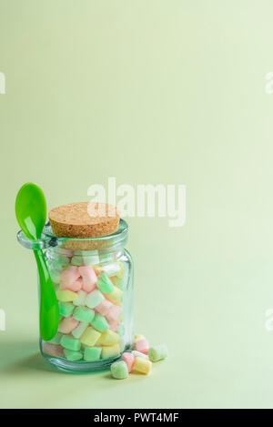 Minimale Bild mit einer Glasflasche voll von verschiedenen farbigen kleinen Marshmallows und eine grüne Löffel befestigt ist, auf ein grünlich Papier Hintergrund. Stockfoto