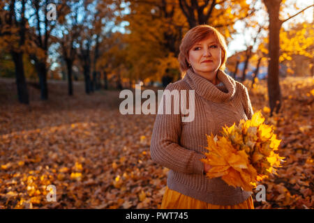 Frau mittleren Alters sammelt Ahornblätter bei Sonnenuntergang. Lady Wandern und Genießen im Herbst Natur tragen stylisches Outfit Stockfoto