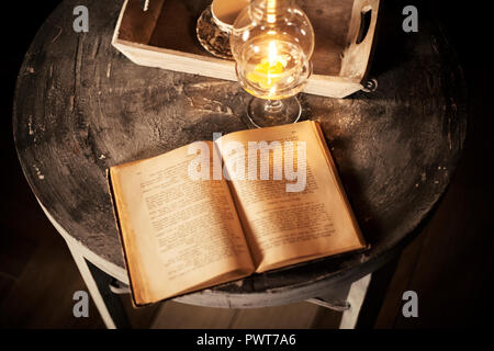 Offenes Buch unter der Lampe mit einer Kerze auf Holztisch, vintage Szene Stockfoto