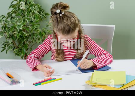 Ein jüngeres Schulmädchen mit Brille schreibt etwas mit der linken Hand auf dem Notebook- und setzt sich an den Tisch. Zurück zum Konzept der Schule. Das Kind ist links Stockfoto