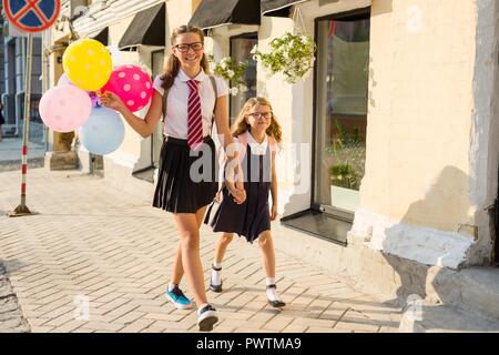 Zwei Mädchen sind zu Fuß entlang der Straße mit bunten Luftballons. Kinder in Schuluniform, Gläser, Rucksack. Stockfoto