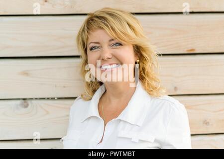 Portrait positive reife Frau mittleren Alters in der Nähe lächelnd. Blonde Frau, outdoor Holz- Hintergrund. Stockfoto
